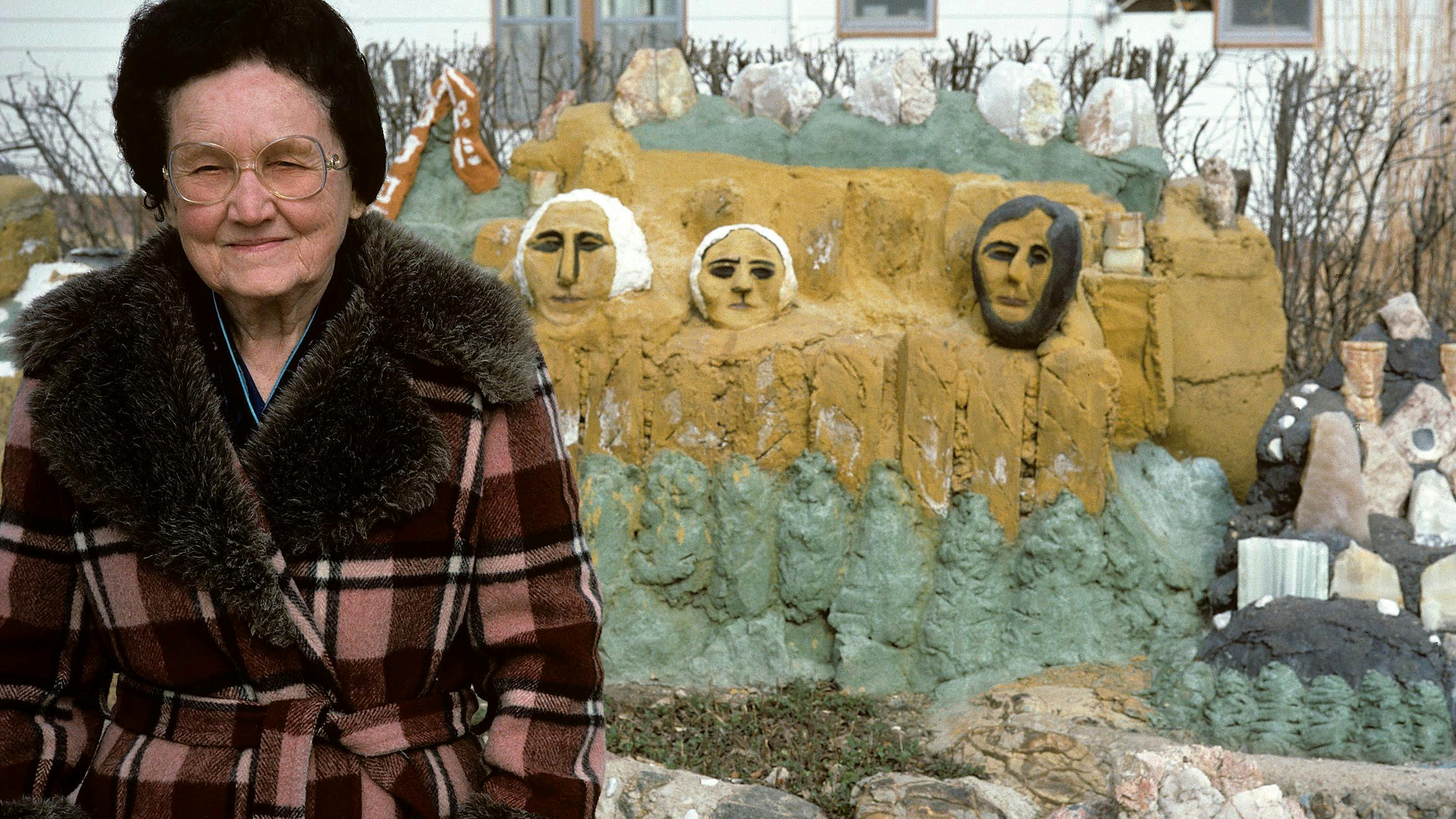 The artist standing in front of her multicolored concrete sculpture of Mt. Rushmore. She is smiling at the camera and is wearing her circular glasses and a brown plaid coat with brown fur trim.