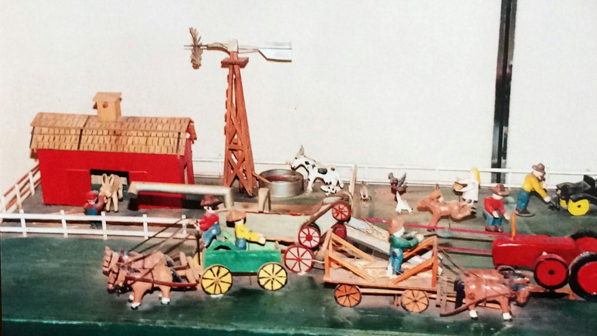 A photograph of a miniature farm including a farmhouse in bright red, cows, horses, farmers, and carriages. The scene depicted shows the miniature farmers doing different tasks around the farm. The objects are reminiscent of children’s toys.